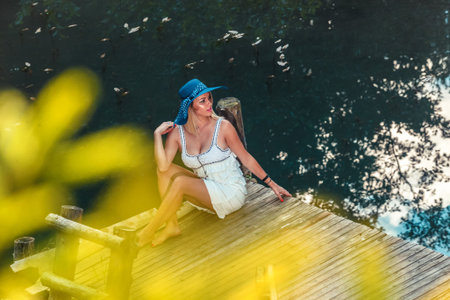 Beautiful blonde girl with a hat on her head and a colorful dress sits on the pier by the water and enjoysの写真素材