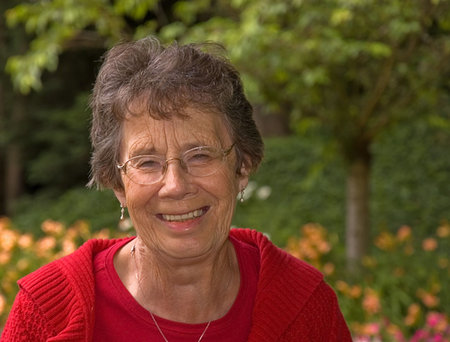 This elderly woman is smiling and laughing with a garden setting in the background, she's wearing a...