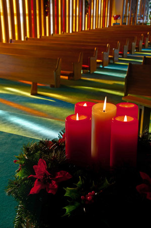 This vertical stock image is lit red and white Christmas candles in an evergreen wreath with holly and Poinsettias with an empty church with rows of wooden pews softly in the background.  Sunlight is shining through stained glass windows.  Beautiful, peacのeditorial素材