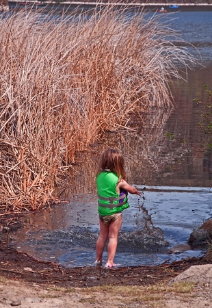 This cute 3 year old Caucasian girl is having great childhood fun  playing in the mud with a mighty splash at the shore of a lake with reeds nearby and fisherman in the background.  Sheのeditorial素材