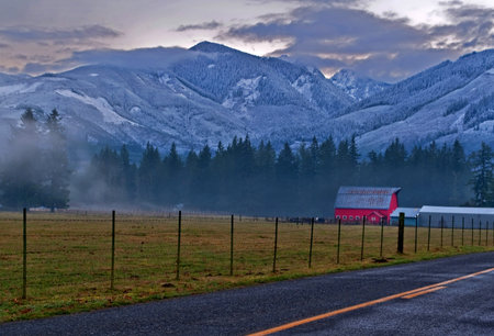 This winter country farm scene has a red barn in the background with a rural lonely road, snowy...