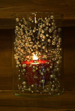 This vertical image is a red votive candle burning in a glass jar filled with beads on a wood shelf   Beautiful, cozy home decor item の写真素材