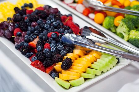 Fruit tray with fresh berries and fruit with metal tongs for serving.  Blueberries, blackberries, strawberries, cantaloupe honeydew melon and grapes.  Vegetables and cheese and blurred in the background.の写真素材