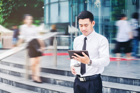 Businessman using a digital tablet office outdoors and Blurred People Walking in Front of Modern Office Buildingの写真素材