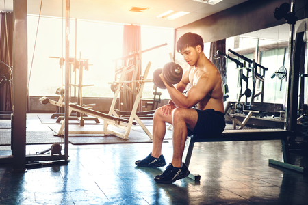 Muscular Man built athlete working out in gym sitting on weightlifting machine and lifting dumbbellの写真素材