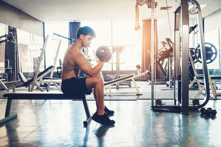 Muscular Man built athlete working out in gym sitting on weightlifting machine and lifting dumbbellの写真素材