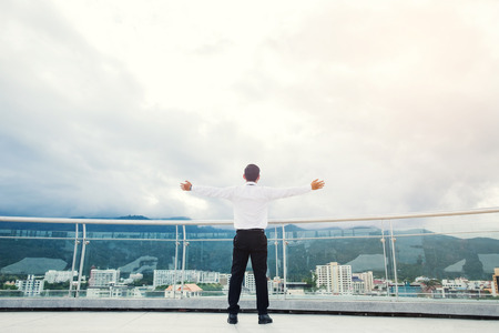 Businessman standing keeping arms raised on a roof and looking at city Success conceptの写真素材