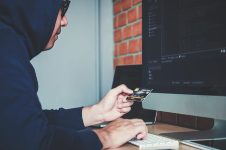 Dangerous Hooded Hacker using credit card typing bad data into computer online system and spreading...