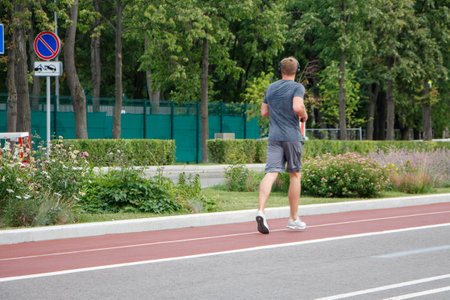 A young man in a gray T-shirt and shorts is running with headphones on a track in a city park.の写真素材