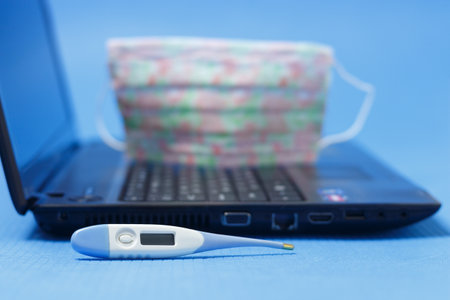 Thermometer on the background of a blurred image of a laptop and a protective mask. Quarantine work concept.の写真素材