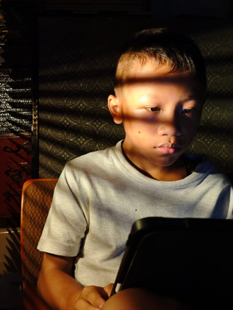 Boy using a tablet computer in a dark room, backlit by sunlight