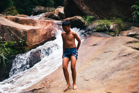 Young Asian boy standing in front of a waterfall in the jungle.の写真素材