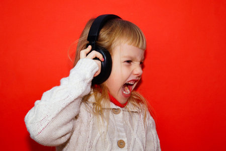 A little girl with headphones, listening to music. A cute girl is listening to music on a red...