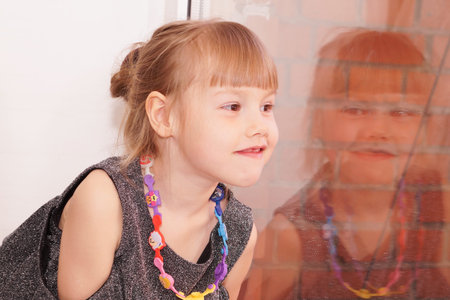 Beautiful little girl with high hairstyle looks out the window. Festive shiny dress of gray color.