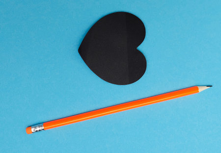 View from above. Orange pencil, black heart-shaped writing paper. Blue background.の写真素材