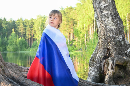 Little charming girl on the shore of the lake in summer at sunset. Wrapped in the Russian flag. Fatherland, patriotism, June 12, Russia Day.の写真素材