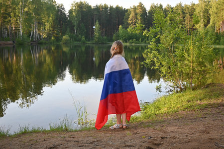Little charming girl on the shore of the lake in summer at sunset. Wrapped in the Russian flag. Fatherland, patriotism, June 12, Russia Day.の写真素材
