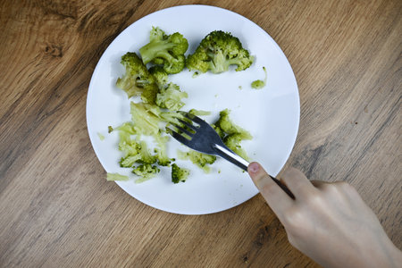 A child eats healthy food with a fork - vegetables, broccoli. White plate on wooden table. A small childs hand.の写真素材