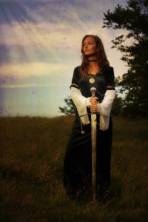 A young beautiful woman with blonde hair in black velvet medieval dress is standing alone on a wild meadow in a mystical midsummer evening light, holding a sword in her handsの写真素材