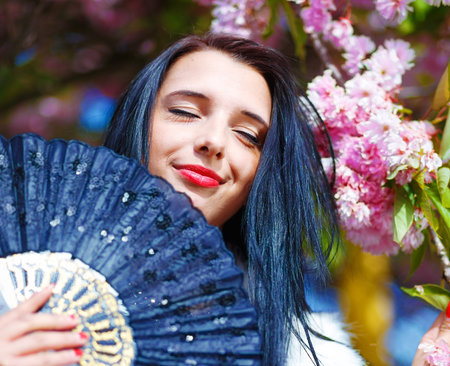 Beautiful Woman with flowers, glamour white fur and black fan in hand, posing next to blooming magical spring rosa sakura flowers. Flower background.の写真素材