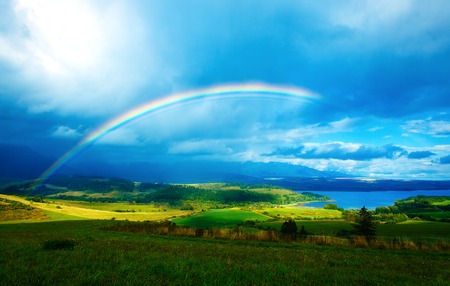 Beautiful landscape, green and yellow meadow and lake with mountain on background with a rainbow in the sky.の写真素材