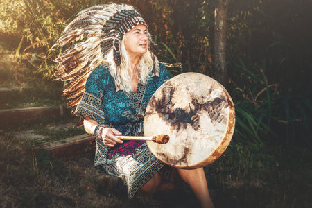 A woman in a Native American headdress sits on grass with a drumの写真素材