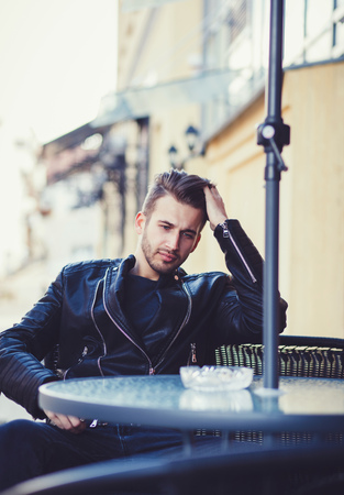 Men`s fashion concept. Stylish man wearing black leather jacket sitting on the streetの写真素材
