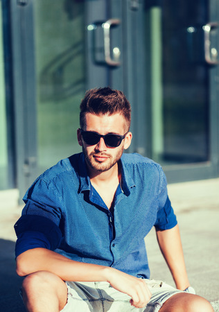 Close up fashion portrait of stylish handsome guy sitting at city wearing blue shirt and sunglasses.の写真素材