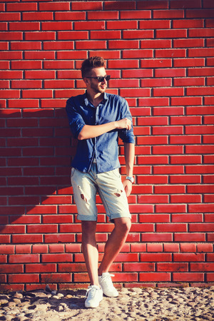 Young man wearing a blue shirt and torn shorts against a brick wallの写真素材