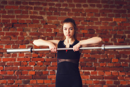 Sporty young woman with barbell on brick background