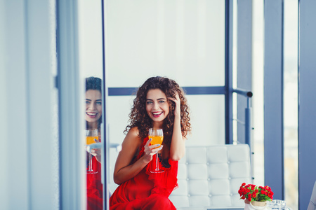 Attractive funny young woman sitting on sofa, holds of fresh juice. Dressed in red dress, with curly hair.の写真素材