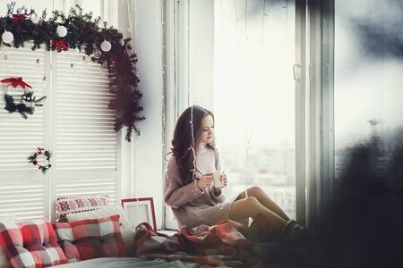 Woman is sitting on the windowsill and drinking hot tea or coffee on christmas dayの写真素材