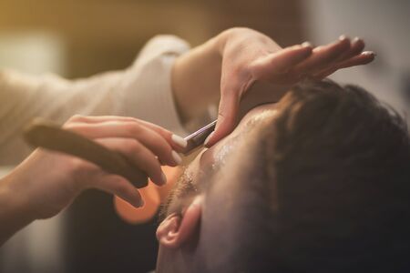 Barber with a dangerous razor shaves a bearded man in a barbershopの写真素材
