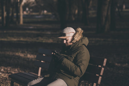 stylish bearded man in a winter jacket sitting on a bench with a smartphone