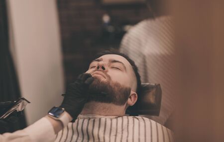 Barber in black gloves takes care of a bearded man in a barbershopの写真素材