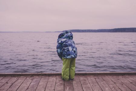 Little boy in a jacket stands on a pier by the seaの写真素材
