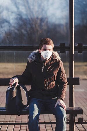 man in a protective mask sits at a bus stop during the pandemic. Epidemic of coronavirus (covid-19)の写真素材