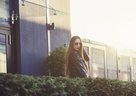 a beautiful girl in a leather jacket and glasses poses on a sunny dayの写真素材