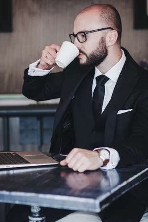 handsome successful man in a business suit drinks coffee and works at a laptop in a cafeの写真素材