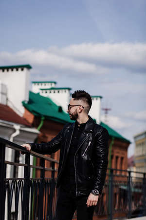 handsome man with a stylish hairstyle in a leather jacket and sunglasses poses against the background of an urban wallの写真素材