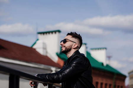 handsome man with a stylish hairstyle in a leather jacket and sunglasses poses against the background of an urban wallの写真素材