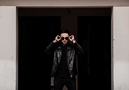 handsome man with a stylish hairstyle in a leather jacket and sunglasses poses against the background of an urban wallの写真素材