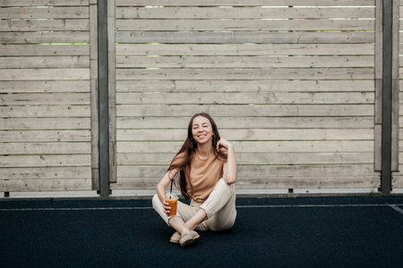 Image of beautiful stylish woman sitting on street with legs crossed on summer day near cup with...
