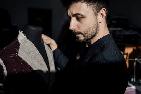 Handsome serious tailor in suit working in atelier with mannequin