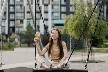 a stylish woman is sitting on a swing and holding a glass of orange juice in her handsの写真素材