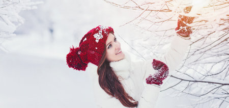 Young woman with long hair in a knitted hat and mittens, a white down jacket, in a winter snowy forest - a park.の写真素材