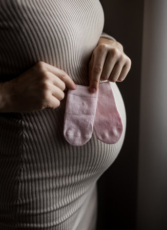 Cropped young pregnant woman wears casual clothes put little tiny socks on belly stay homeの写真素材