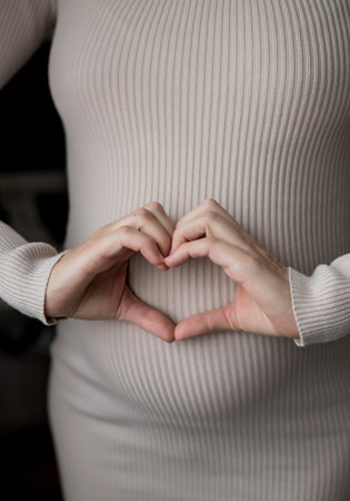 Hands of pregnant woman dressed in comfort home dress and making a heart symbol on her belly. Pregnancy healthy motherhood concept.の写真素材