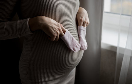 Pregnancy Concept. Pregnant woman showing cute pink baby socks and belly with baby bump motherhood and maternity concept, waiting for childbirthの写真素材