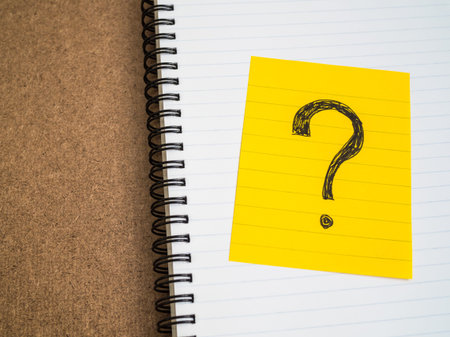 Question mark symbol on yellow sticky note on white spiral notebook on brown board - business...
